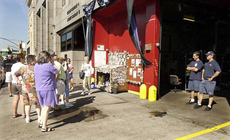 Tourists have made pilgrimages to New York City firehouses since 9/11. Here, FDNY firefighters Scott Gaboff and Eric Taddeo stand patiently as they are photographed by tourists outside the South Street firehouse. Along the wall is a memorial bearing police and fire patches from around the world. This was temporary home to Ladder 10 members while the TenHouse was being rebuilt.  Photo by Mike De Sisti 8/10/02 © 2002 Appleton Post-Crescent